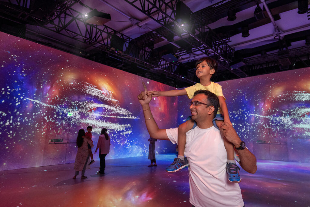 A father carries his son on his shoulders as he walks through an immersive projection experience.