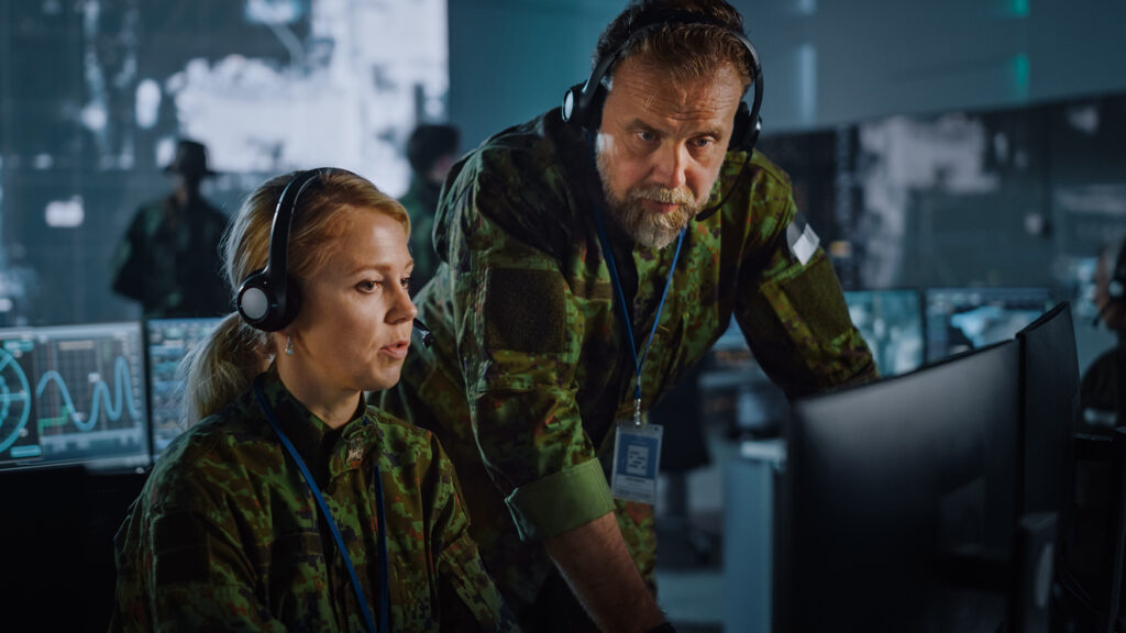 Two military personnel looking at a desktop monitor with a command center video wall behind them.