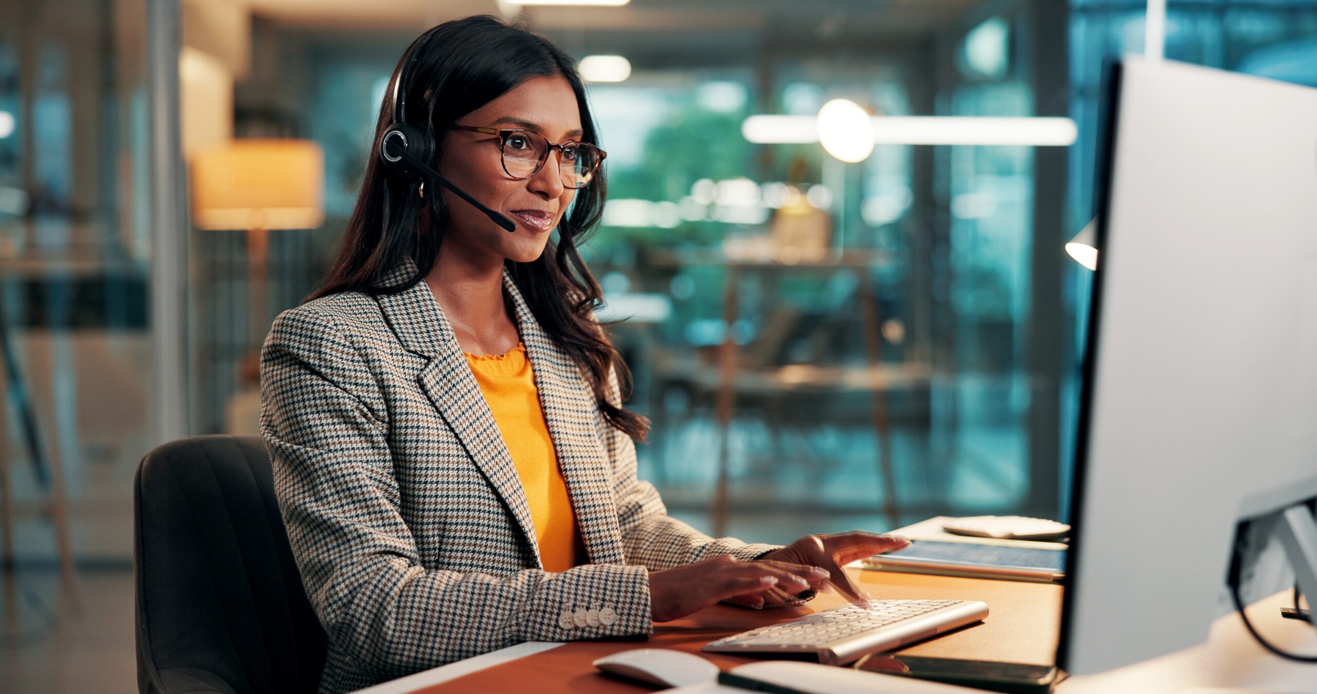 A woman using a headset at a desktop in a modern office, participating in a call and supporting clear, focused communication in a hybrid work environment