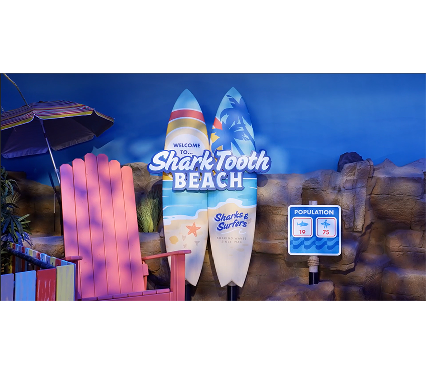 Two surfboards lean against a wall with “Welcome to… Shark Tooth Beach” written across the boards.